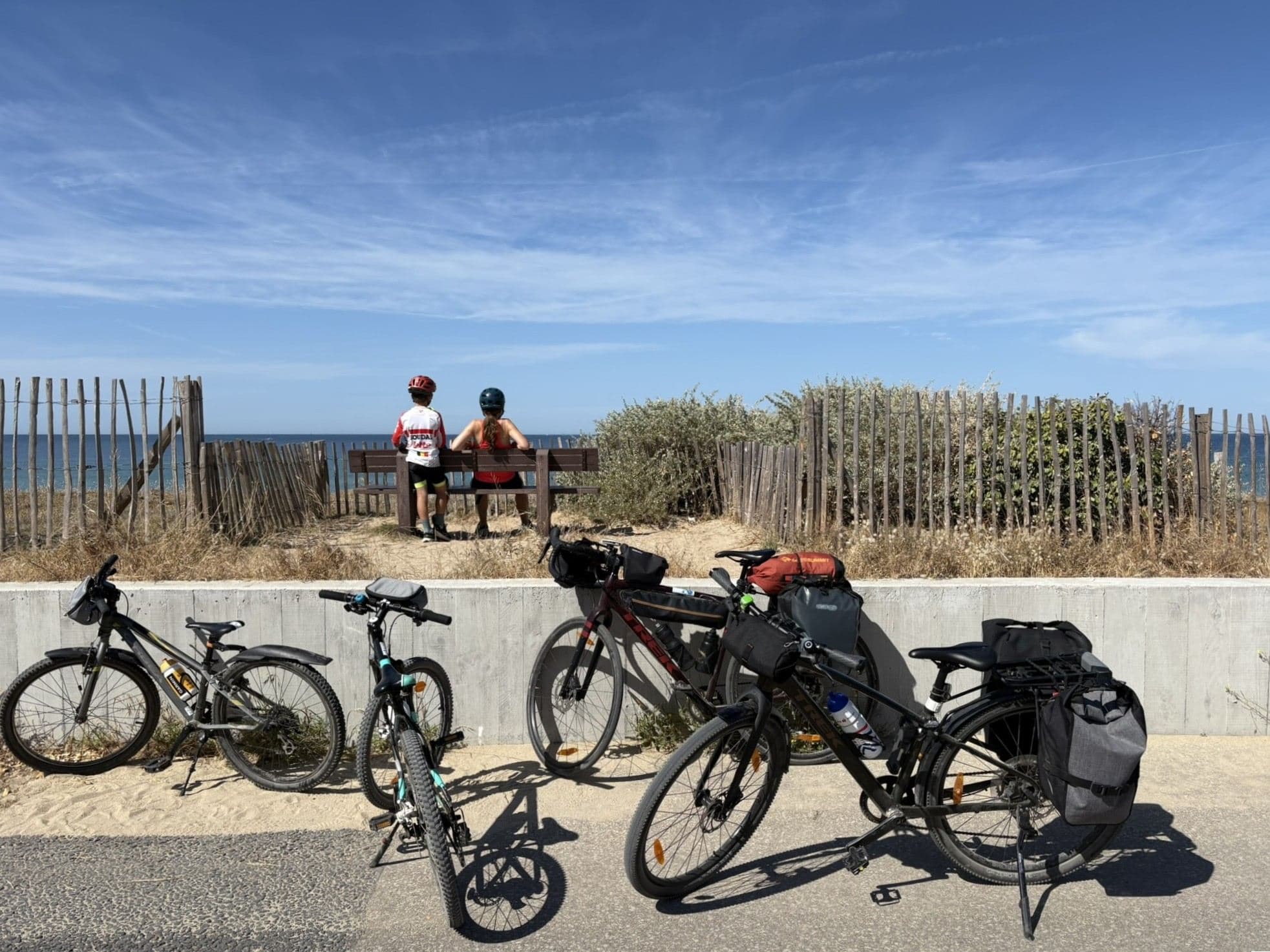 Vier fietsen met fietstassen tegen een muurtje, twee fietsers van achteren met zicht op zee.